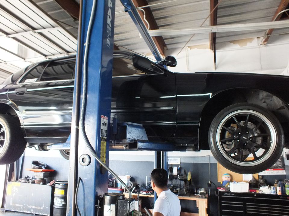 Black sedan raised on a blue two post lift with technician working below in repair shop