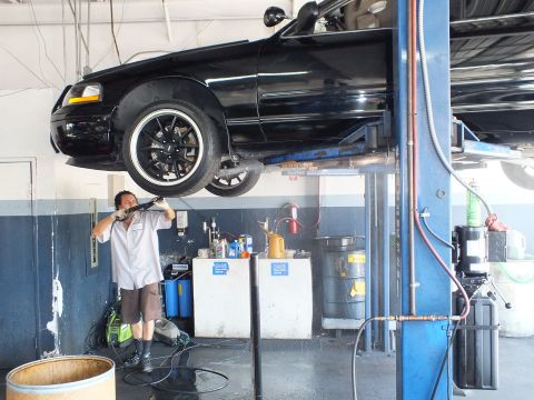 2003 Ford Crown Victoria - Technician pressure-cleaning under a black sedan lifted overhead on shop hoist.