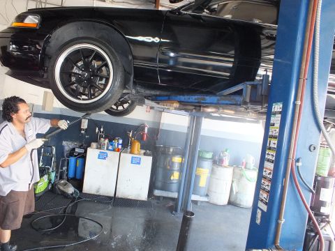 2003 Ford Crown Victoria - Mechanic spraying the undercarriage of a hoisted black sedan, front wheel and fender visible.