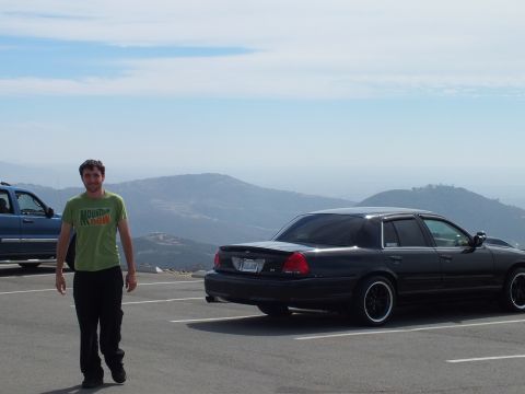 2003 Ford Crown Victoria - Person posing in an overlook parking lot next to a black four-door sedan, hills in the distance.