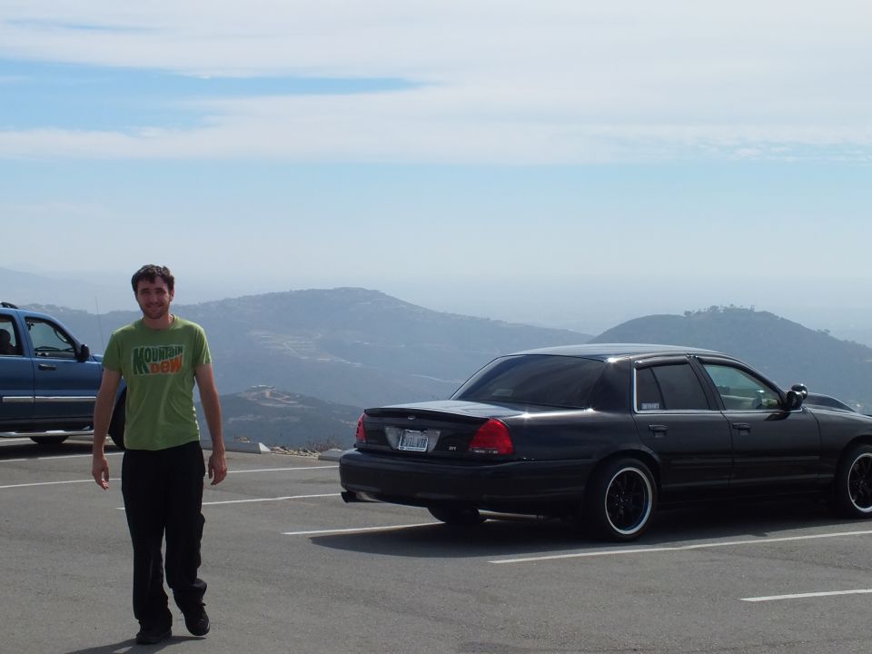 Man in green shirt standing in a hilltop parking lot beside a black sedan with mountain view