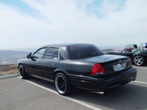 2003 Ford Crown Victoria - Black four-door Ford sedan on staggered black wheels at a hilltop lot, roadster in background.