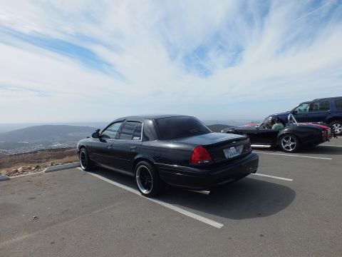 2003 Ford Crown Victoria - Black four-door sedan on dark wheels at a hilltop overlook, SUV and red roadster behind.