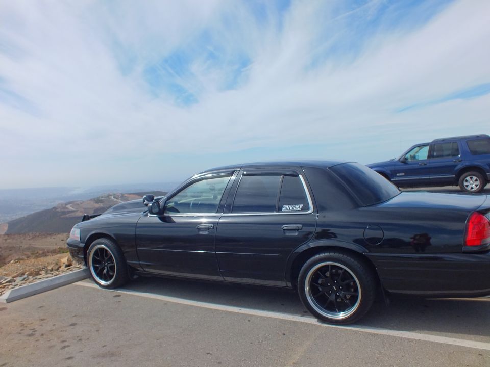 Side profile of a black Ford sedan with DRIVE-BY decal on black wheels beside an SUV at an overlook