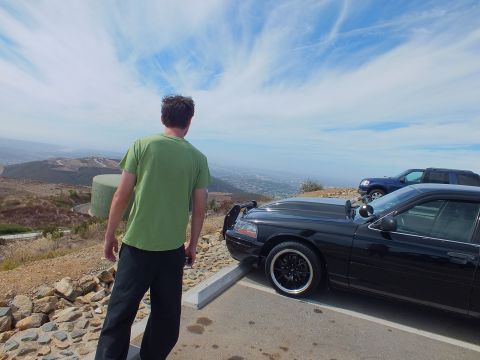 2003 Ford Crown Victoria - Person looks out over a hazy valley beside a black sedan parked at a mountaintop lot.