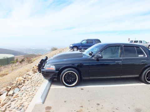 2003 Ford Crown Victoria - Lowered black sedan on black wheels sits in a paved lot with a blue SUV behind.