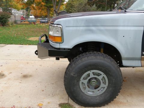 1989 Ford Bronco - The front fender and wheel of a light-colored truck are visible, an aftermarket bumper and off-road tire.