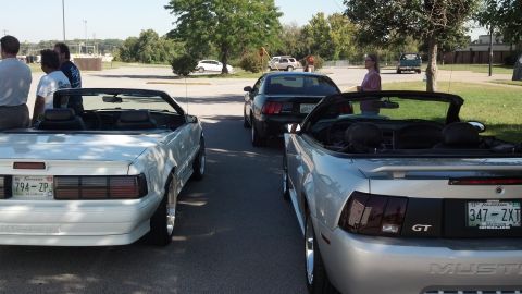2007 Ford Mustang - Several Ford Mustangs are gathered in a parking lot on a sunny day, with people socializing near one of the vehicles.