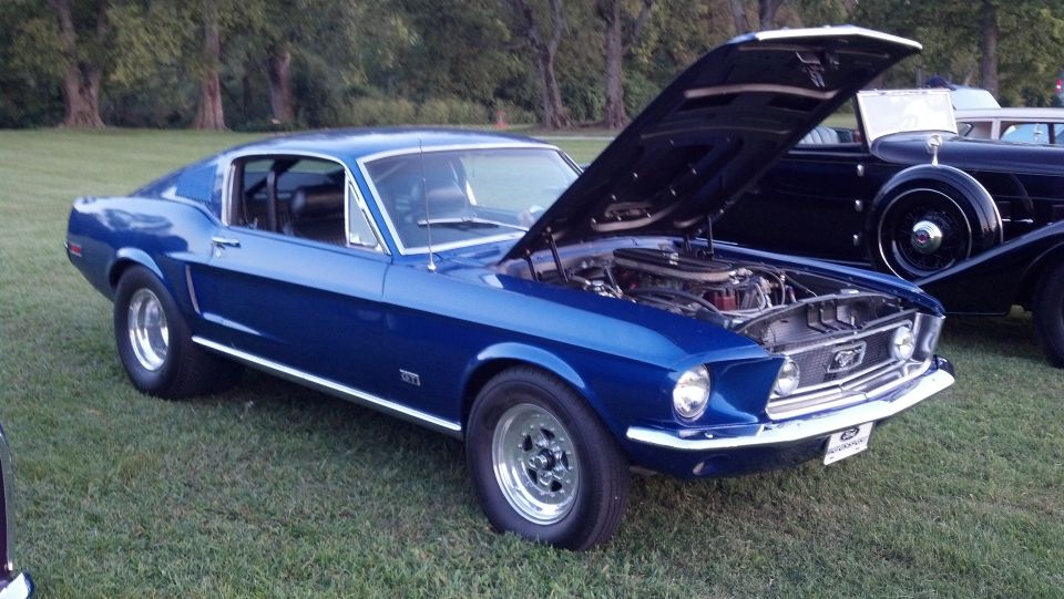 Blue classic Ford Mustang fastback with hood open, showing engine. Another classic car is parked to the right