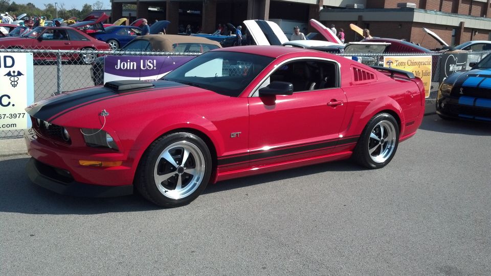 Red 2007 Ford Mustang GT with black racing stripes and aftermarket wheels parked at a car show