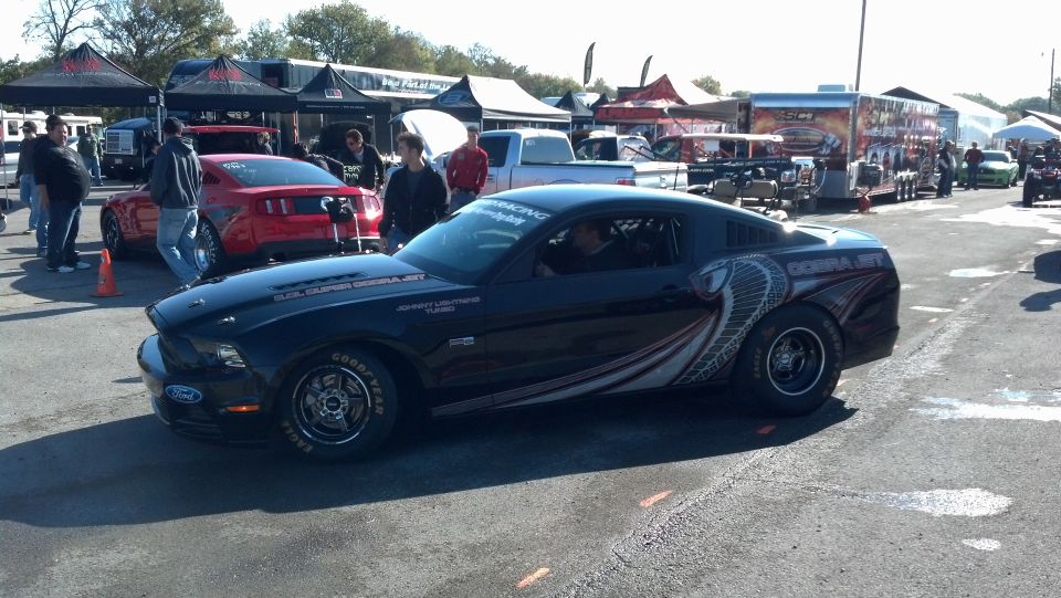 Black drag racing Mustang with cobra graphics on the side, parked at a track with tents and other cars