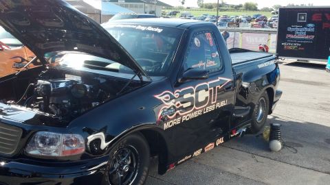 2007 Ford Mustang - Modified black pickup truck with racing decals and an open hood at a drag strip event.