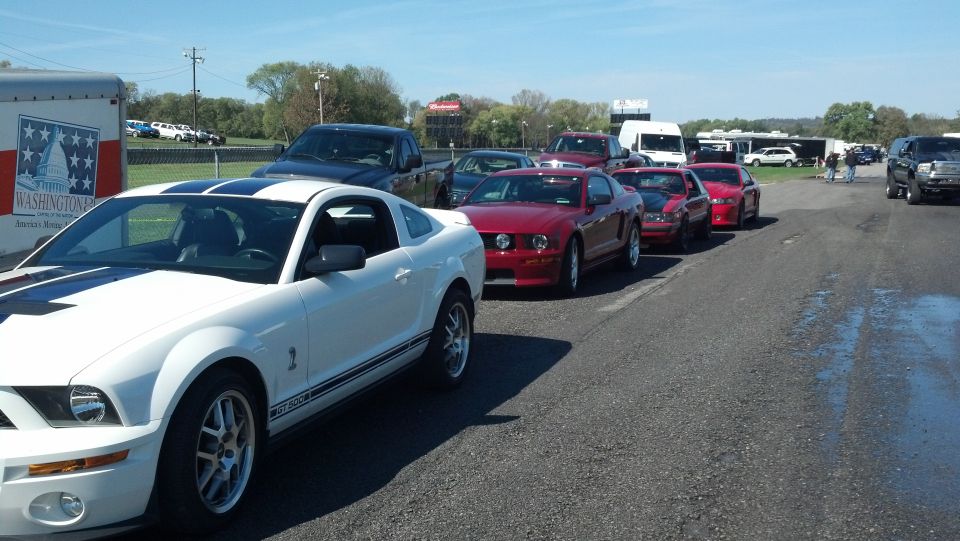 White Ford Mustang in foreground, line of red Mustangs behind it, trailer on left