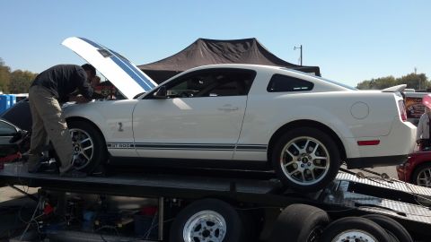 2007 Ford Mustang - A white 2007 Ford Mustang GT500 is loaded onto a trailer while a person works under the open hood.