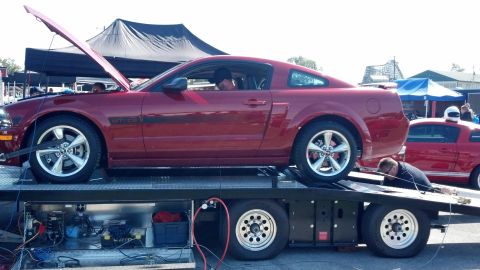 2007 Ford Mustang - A red 2007 Ford Mustang GT is loaded onto a trailer at an event, with its hood up and another Mustang visible in the background.