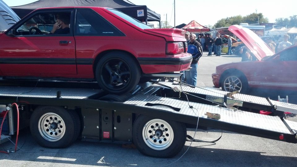 Red coupe on trailer, side view. Another red car with hood up nearby