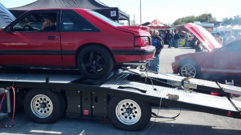 2007 Ford Mustang - Two red coupes are on a car trailer at an outdoor event with spectators.