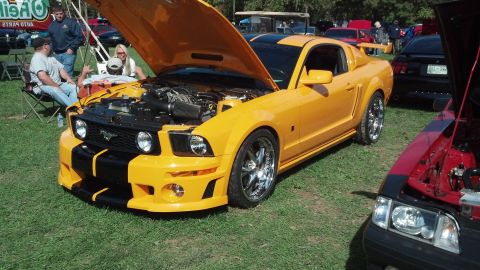 2007 Ford Mustang - A yellow 2007 Ford Mustang with racing stripes is displayed with its hood open at an outdoor car show.
