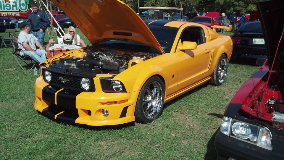 Yellow 2007 Ford Mustang with black racing stripes, hood open, parked on grass next to other cars