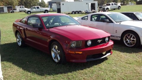2007 Ford Mustang - A red Ford Mustang is parked on a grassy field with other vehicles and trailers visible in the background.