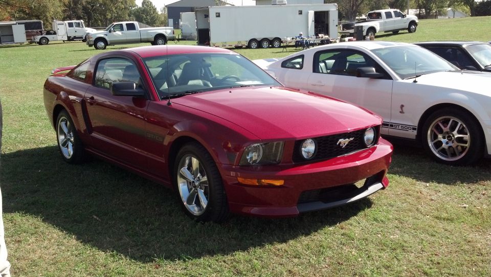 Red Ford Mustang parked on grass next to a white Mustang. Trucks and trailers in background
