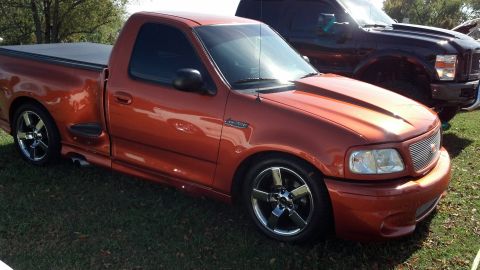 2007 Ford Mustang - This orange Ford F-150 Lightning pickup truck features custom wheels and a hood scoop, parked next to another truck.