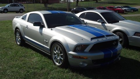 2007 Ford Mustang - A silver 2007 Ford Mustang Shelby GT500 with blue racing stripes is parked on a grassy area next to another Mustang.