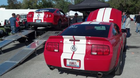 2007 Ford Mustang - Two red Ford Mustangs with white racing stripes are parked at an outdoor event, with one loaded onto a car trailer.
