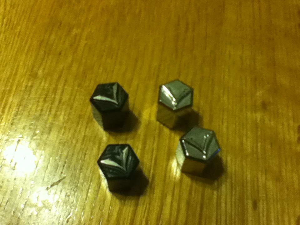 Four lug nuts, two black with silver accents, two chrome, on a wooden surface