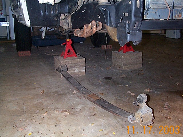 Leaf spring removed on garage floor beneath vehicle raised on red jack stands and wood blocks