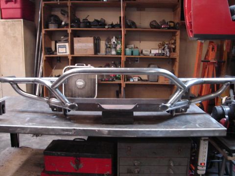 1989 Ford Bronco - A finished bare-steel tube front bumper rests on a steel welding table in a home garage, with a center hoop, wraparound tube ends, a flat l…