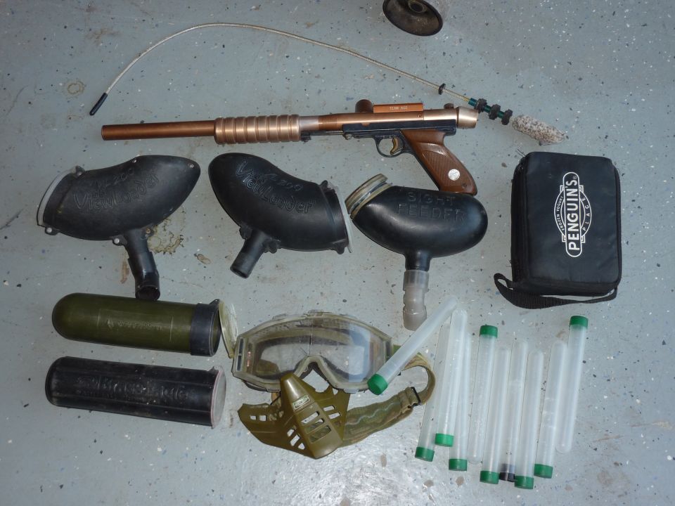 Paintball gun, hoppers, mask, goggles, and tubes laid out on a concrete surface