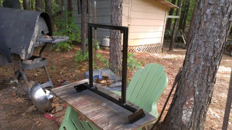 1978 Ford Bronco - An unassembled metal frame sits on a table in a wooded area, with a barbecue grill and a chair nearby.