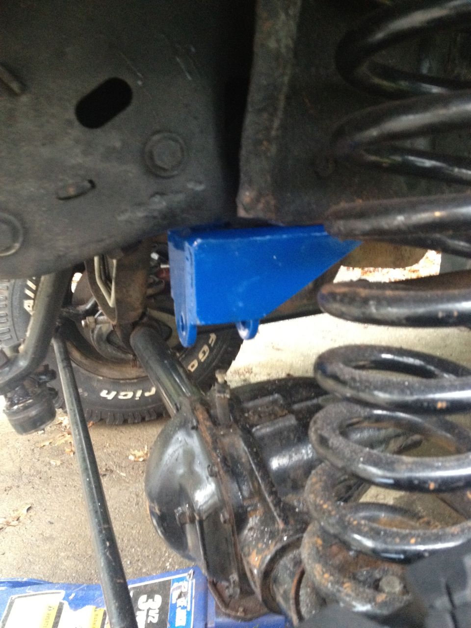 Blue suspension bracket mounted to a vehicle's front axle assembly, near a coil spring