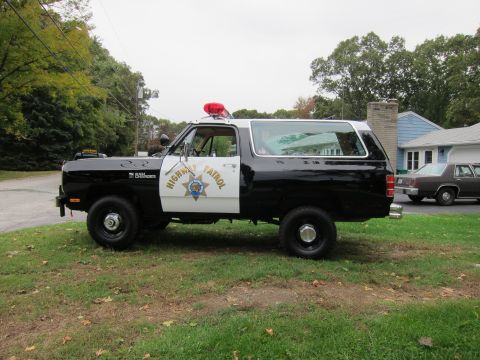 1987 Dodge 1500 - A black and white police SUV, identified as a Ramcharger, is parked on a grassy area next to a house and another car.