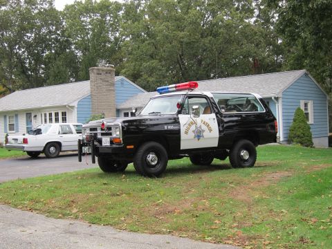 1987 Dodge 1500 - This highway patrol SUV, likely from the 1980s, is parked in a residential driveway next to a white sedan.