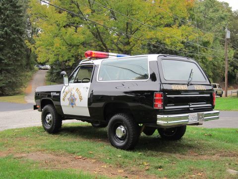 1987 Dodge 1500 - This black and white police SUV, likely from the 1980s, is parked on a grassy area with trees in the background.