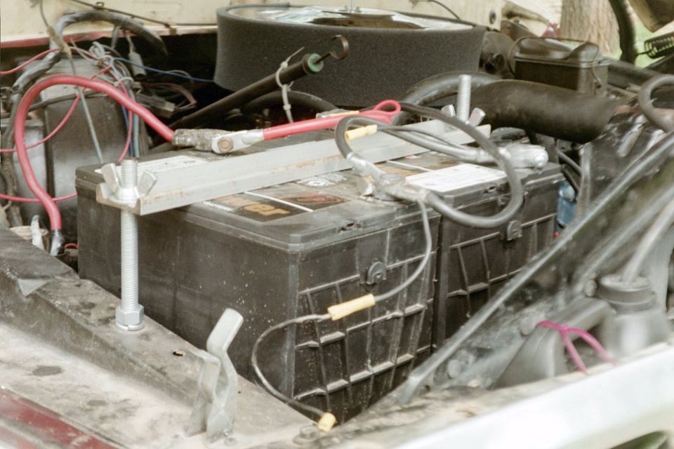 Dual batteries strapped down in an engine bay with red cables and aftermarket hold-down bar