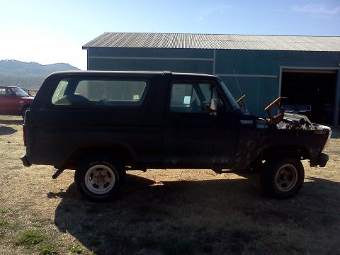 1978 Ford Bronco - Side view of a black Ford Bronco with its hood up, parked in a dry, grassy area near a building.