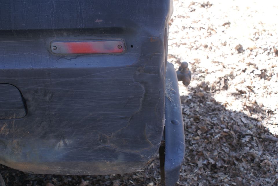 Dark rear bumper with a red reflector and a tow hitch, showing scratches and damage