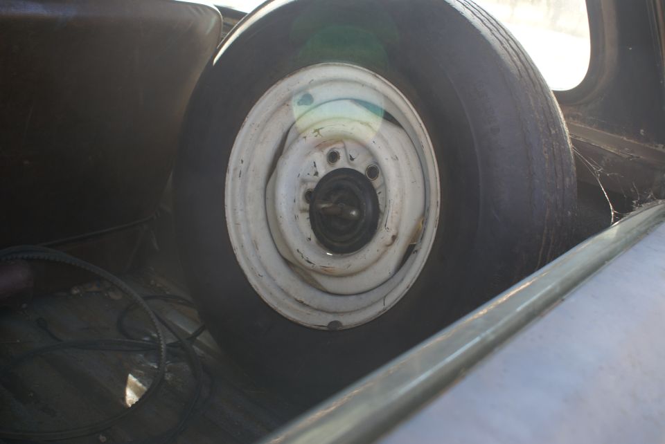 Spare tire mounted inside a vehicle, showing a white rim and black tire. Cobwebs are visible in the background