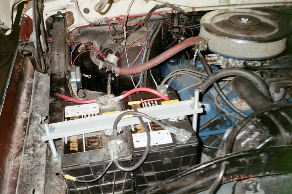 Engine bay with blue Ford engine, dual batteries on a hold-down bar, red cables and belt-driven fan