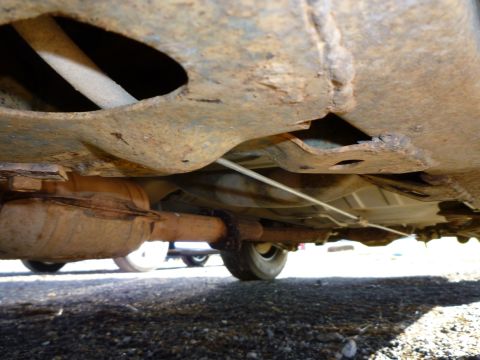1997 Ford Crown Victoria - The underside of a heavily rusted vehicle, showing the exhaust system and frame, with another car partially visible in the distance.