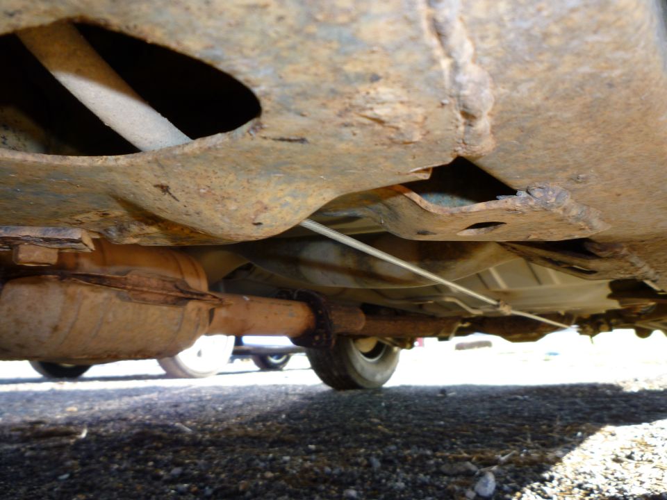 Underside view of rusty exhaust system and frame with a visible tire and another vehicle in the background