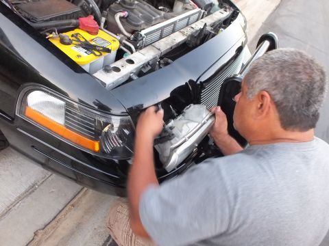 2003 Ford Crown Victoria - Person fits a headlight assembly into a black car beside a yellow-top battery.