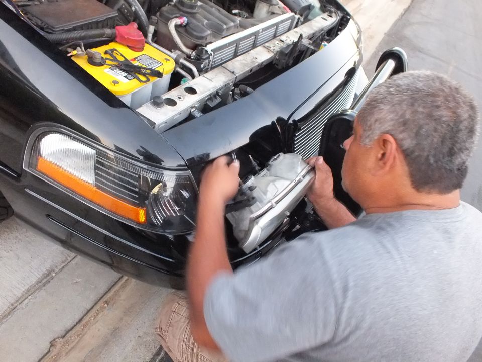 Man installing headlight on black sedan with yellow Optima battery visible