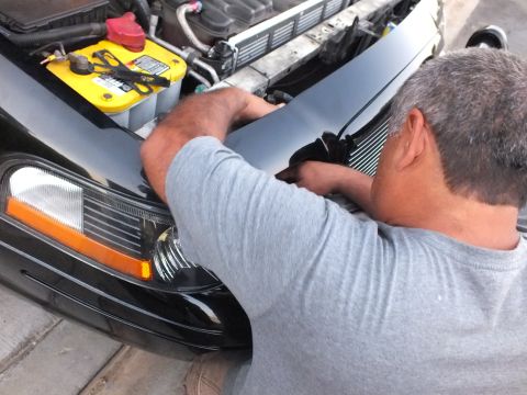 2003 Ford Crown Victoria - Person positions a dark plastic panel on the front of a car with hood raised.