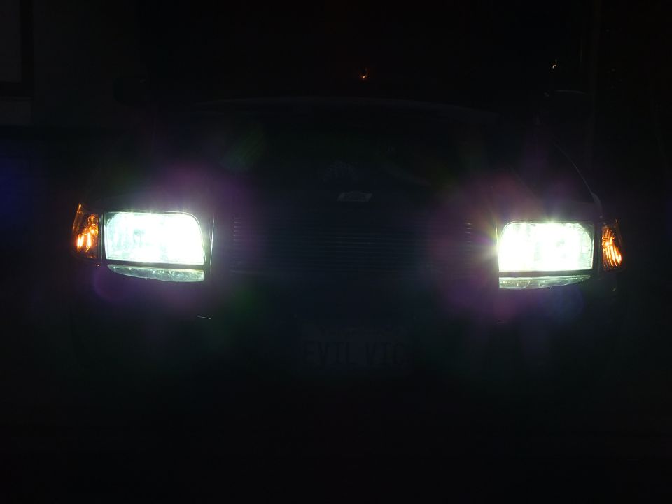 Dark front view of black Crown Victoria with two bright HID headlights and faint EVIL VIC plate