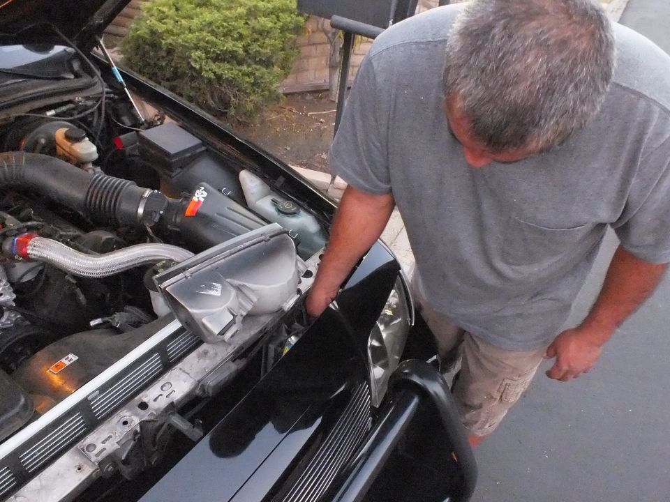 Man in gray shirt leaning over open engine bay of black Crown Victoria in a driveway