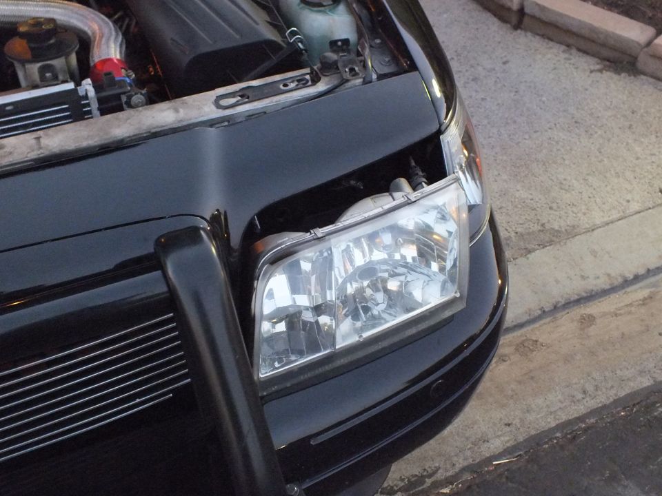 Clear projector-style headlight assembly mounted on black Crown Victoria with push bar at night